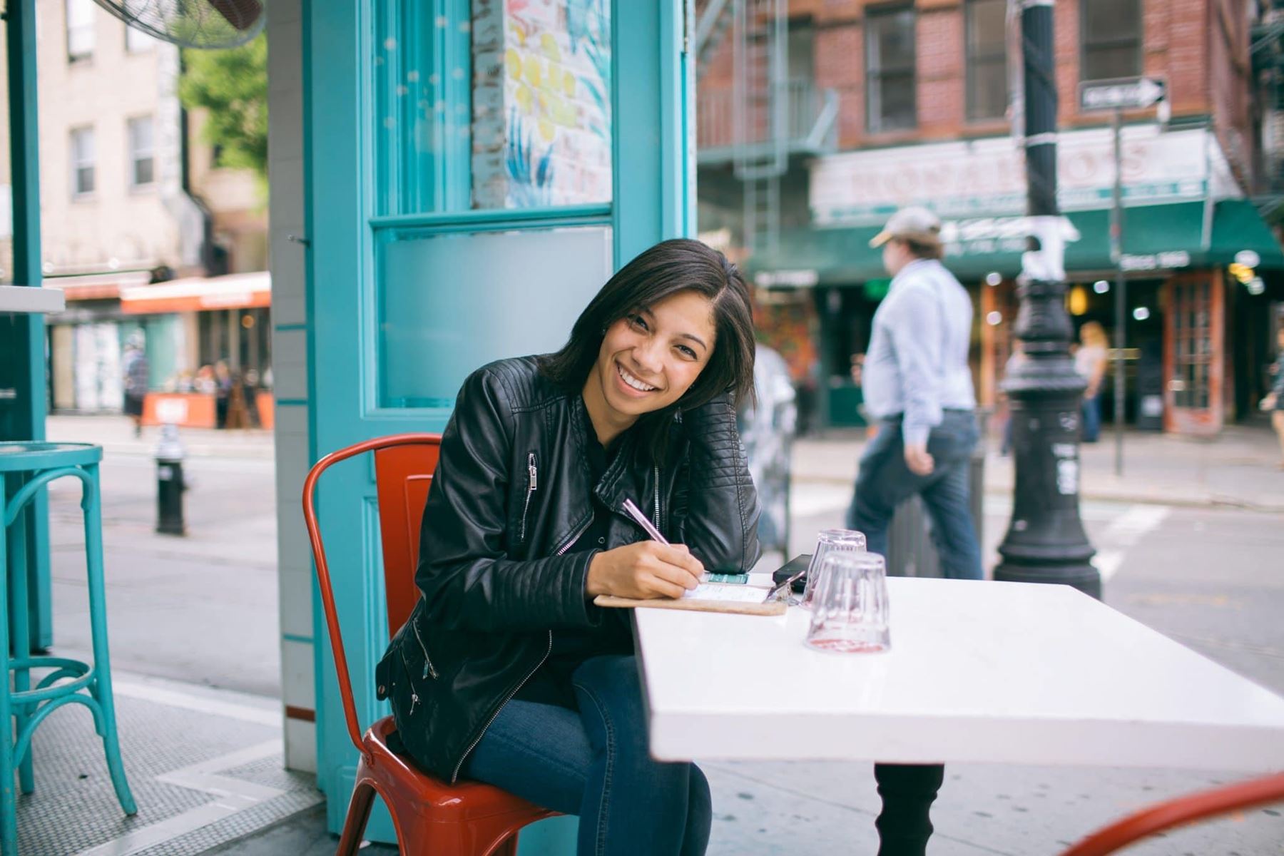 Woman sitting at an out door diner, smiling and writing on a notepad